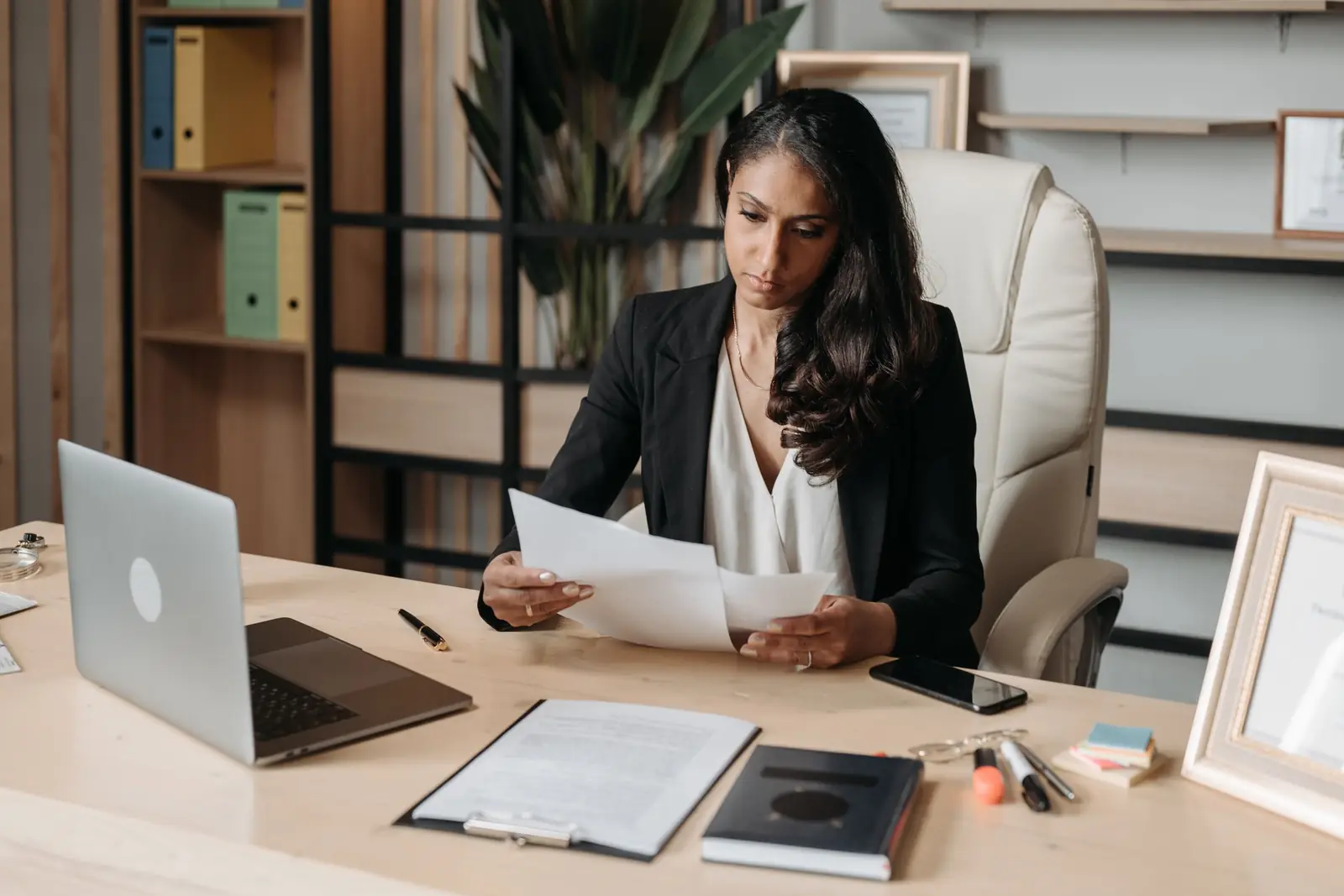 A woman reviews printed documents at her desk beside an open laptop.