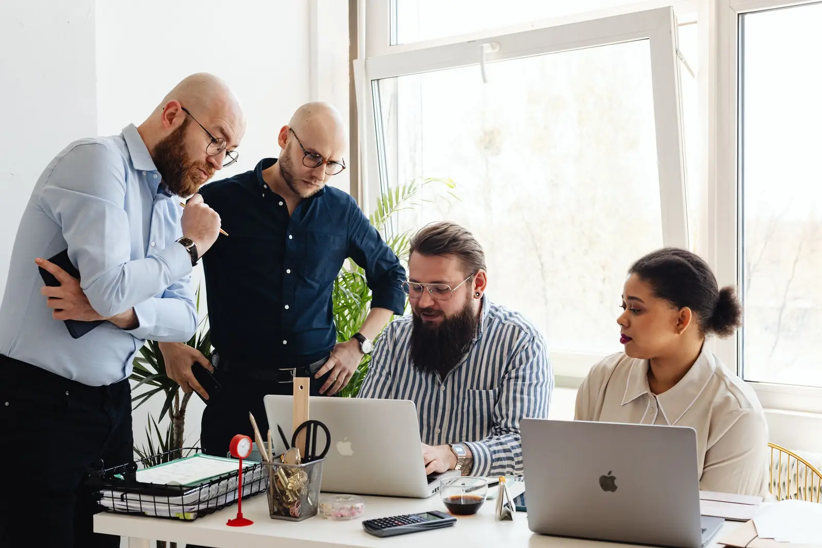 Four colleagues stand around a meeting table looking at a laptop together, printed analyses spread out beside it.