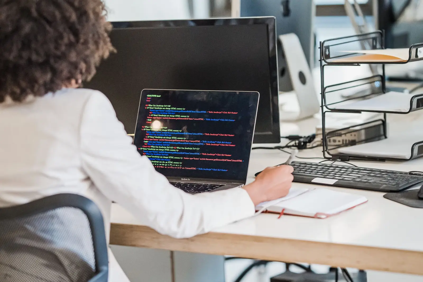 A developer works at a laptop with code on screen, at a desk in an open-plan office.